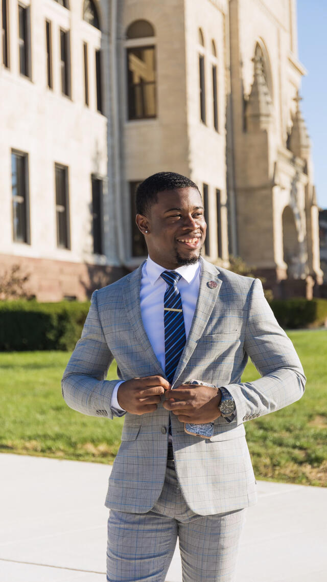 a Bahamian man buttoning his suit jacket wearing a gray suit white shirt and blue tie