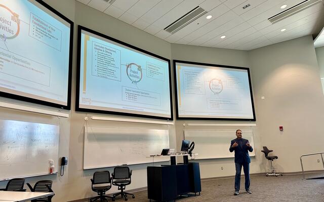 person stands in front of three large television screens on the wall in a lecture hall