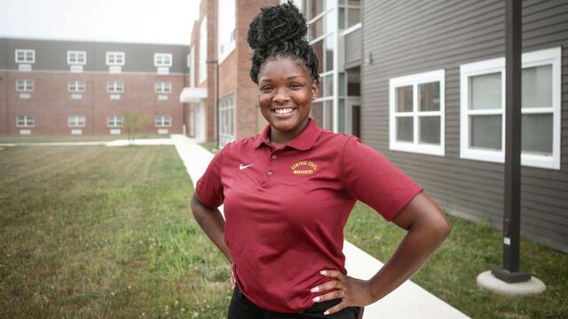 A young Black woman in maroon stands with hands on hips in front of residence halls at Central State University