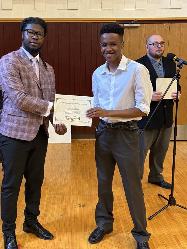 an african american man with locs wearing a plaid suit jacket and black pants presents a certificate to a young black man in a white button up shirt and slacks