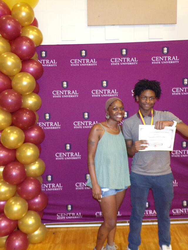 a high school student and parent holding a certificate with maroon and gold balloons nearby